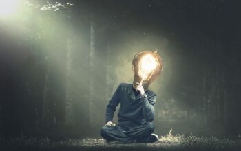 a drably dressed person with a lit light bulb head sits thinking on a misty moody forest floor