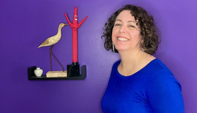 portrait of Adrienne Montgomerie with curly hair down to their shoulders, smiling in front of a bright purple wall beside a bird carving and an "air dancer" failing armed tube man